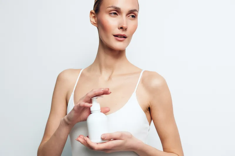 Young woman with clear skin holding white cosmetic bottle, skincare routine concept, natural beauty, minimal makeup, white background, clean fresh look, wellness focus