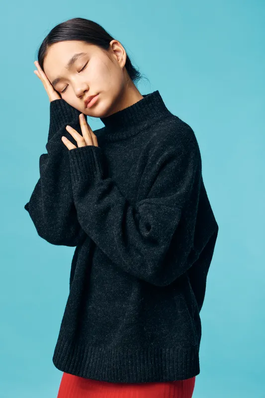 Woman wearing black sweater resting her head on hand with eyes closed against blue background. Calm, peaceful expression in casual cozy clothing for autumn or winter season.