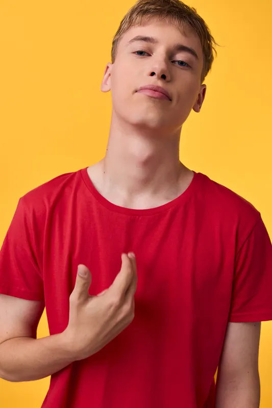 Young man in a red shirt stands against a yellow background with a confident pose and a subtle hand gesture on the chest, conveying bold, modern energy suitable for fashion and lifestyle visuals