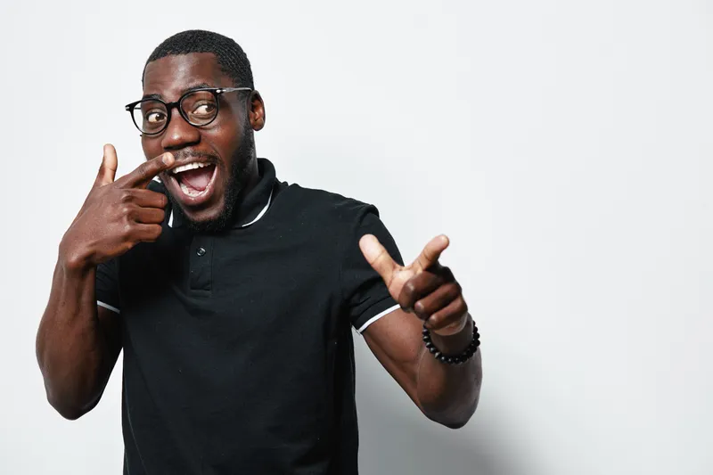 African American man with dark skin and glasses pointing at his smile with finger, wearing black polo shirt, expressing happiness and confidence on white background.