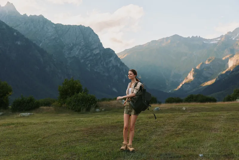 Young woman with backpack walks across a grassy valley beneath dramatic mountains, enjoying the fresh air and open landscape