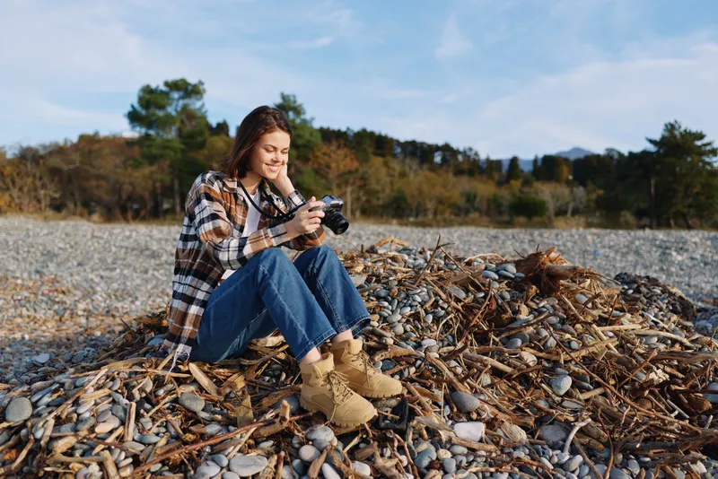Woman photographer sits on a rocky beach with a camera, wearing a plaid shirt, jeans and boots, capturing outdoor nature and candid moments by the pebbled shore.
