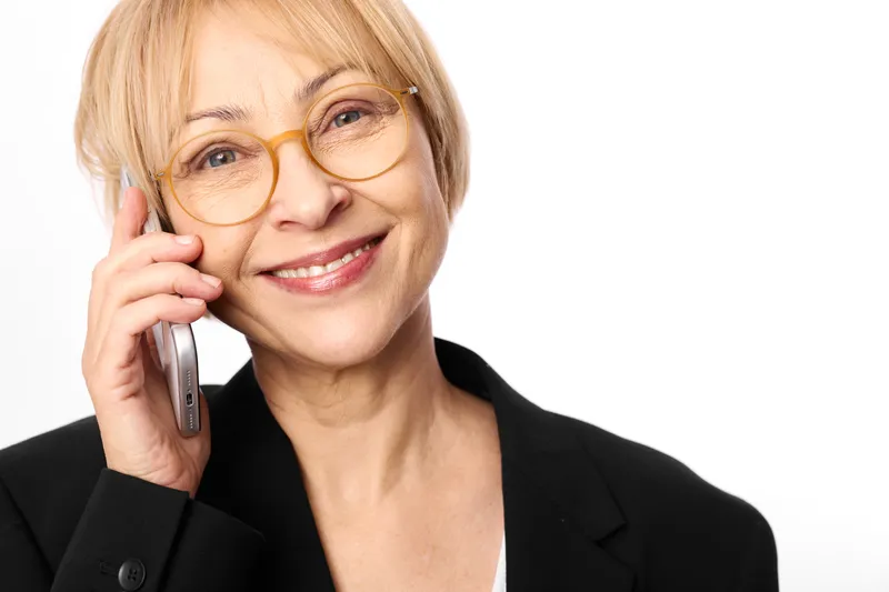 Smiling senior woman wearing glasses holding a smartphone to her ear, studio portrait on white background, communication and business call, calm confident mood, soft light