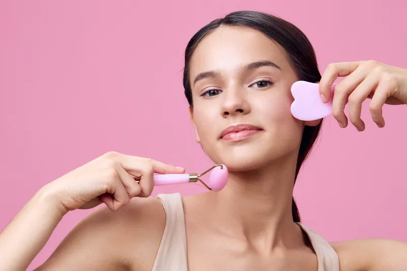 Young woman using a pink facial roller and beauty sponge on a pastel pink background, promoting skincare and wellness concepts with a serene expression