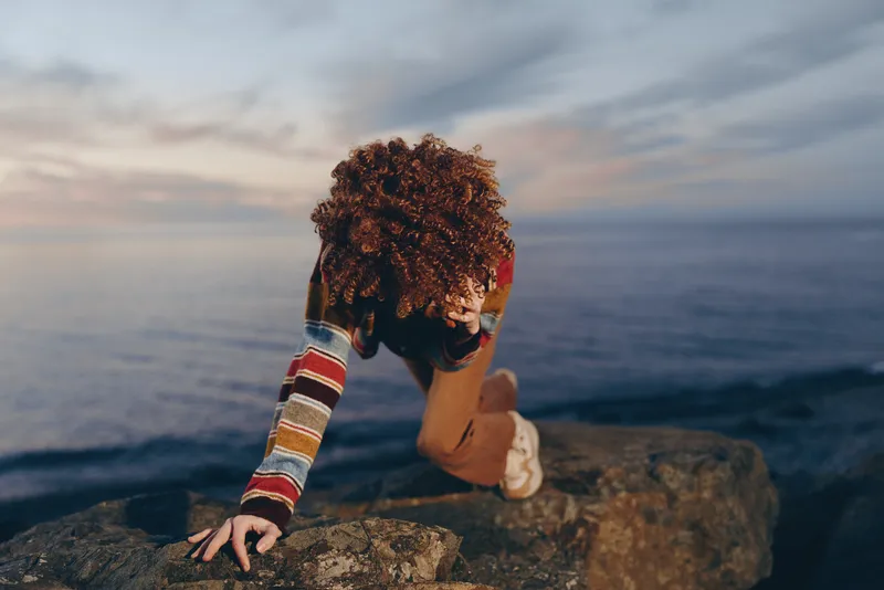Woman lifestyle outdoors on a rocky beach at sunset, curly hair flowing, leaning forward with hands on the cliff, ocean and horizon in soft twilight, natural light, calm ocean waves