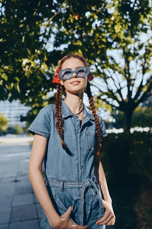 lifestyle woman in modern boho-western denim outfit posing outdoors with braided hair and oversized sunglasses, film color street style fashion bright summer day