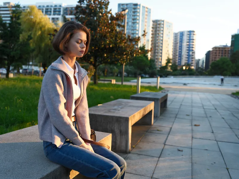 A focused professional woman sits on a park bench in a modern urban setting, checking her phone with a gentle smile, creating a calm, confident workday moment.