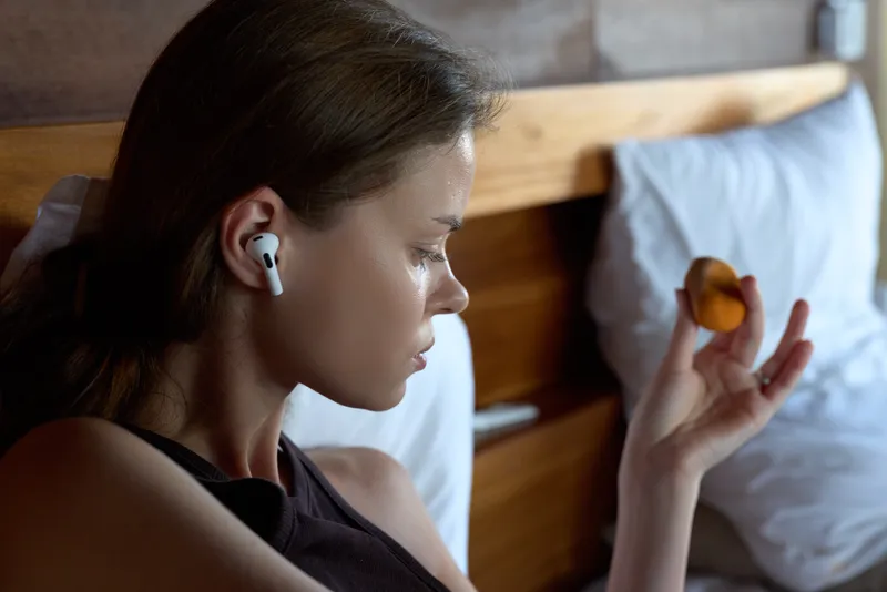 Calm woman in bed wearing wireless earbuds, holding a small fruit near her fingers, focused on a quiet morning moment, soft light, intimate lifestyle scene.