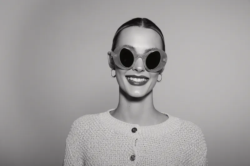 Smiling Caucasian woman with fair skin wearing round sunglasses and earrings poses against plain background. Concept for fashion, eyewear, and happy mood.