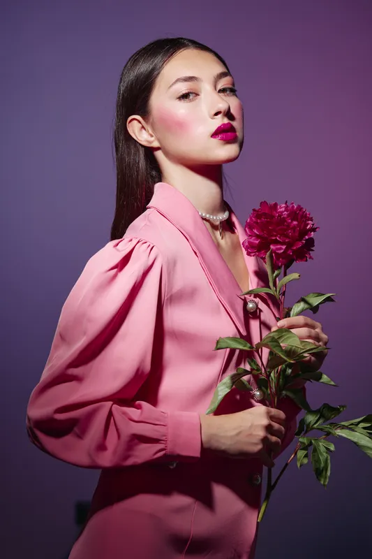 Young woman in a pink blouse holds a deep pink flower with green leaves. Her makeup features bold pink lips and highlight. Studio portrait with strong vibrant beauty and fashion concept.