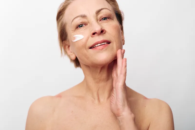 Mature woman with short hair applies cream on her face with gentle smile, isolated on white background. Concept of skincare, aging beauty, self-care, and healthy skin routine for women.