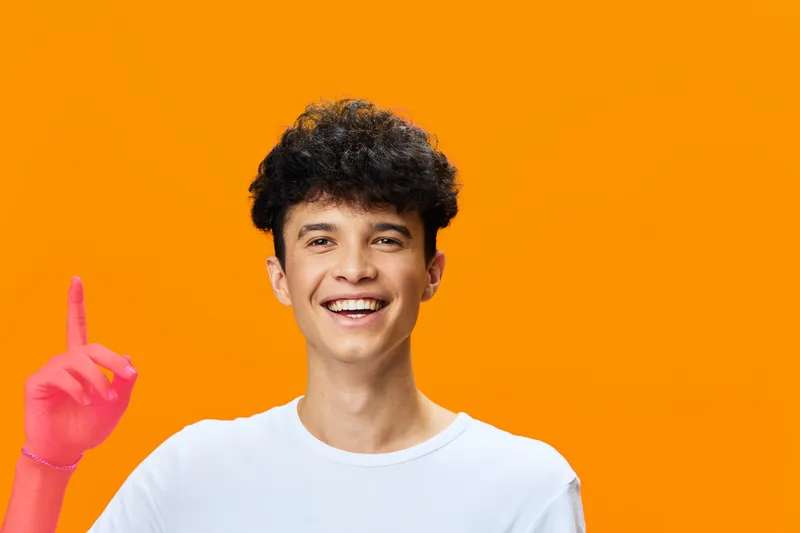 smiling young man pointing finger up against bright orange background, casual white t-shirt, curly dark hair, happy expression, studio portrait