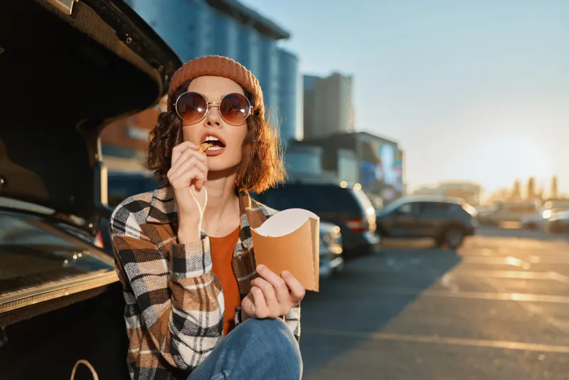 Woman car food parking sunglasses beanie casual urban scene of a candid young woman sitting at an open trunk eating takeaway, authenticity and golden hour glow, mindful living and emotional
