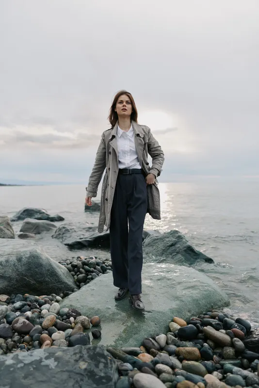 Woman in long coat and white shirt walks on rocks by the sea shore under cloudy sky. Casual fashion concept with natural landscape and moody atmosphere.