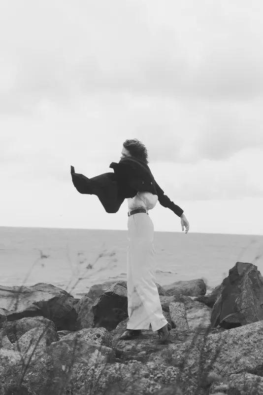 Windblown figure stands on rocky shoreline facing the sea, scarf trailing in the breeze, wide coat edges fluttering, calm horizon, moody monochrome scene capturing solitude resilience and freedom in