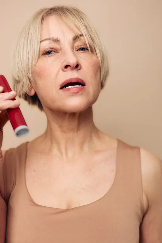 Mature woman with short blonde hair applying deodorant underarm in beige tank top. Close-up portrait on light background. Concept of personal hygiene, skincare routine, and self-care.