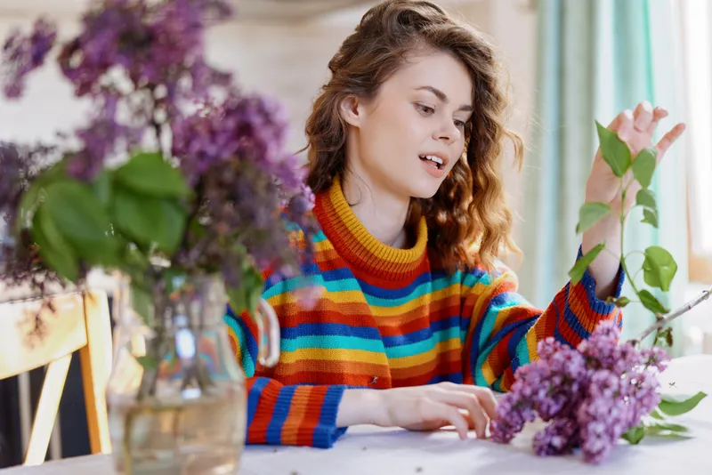 A woman in a colorful striped sweater arranges lilac flowers at a bright home table, capturing a calm, creative moment of daily life and floral care in a cozy domestic setting.