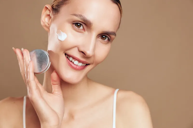 Cheerful woman applying cream to her cheek with a round pad, skincare moment, studio portrait, soft lighting, neutral background, fresh complexion, natural colors, relaxed mood