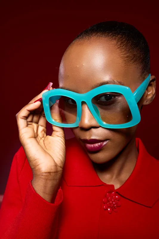 Close-up portrait of a dark skinned African woman wearing large turquoise glasses and red clothing. She poses confidently on a red background. Concept of fashion, beauty, and style.