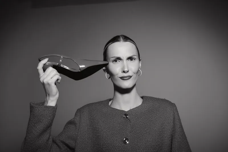 Portrait of a white caucasian woman with fair skin holding a high heel shoe near her head with a neutral expression in studio lighting.