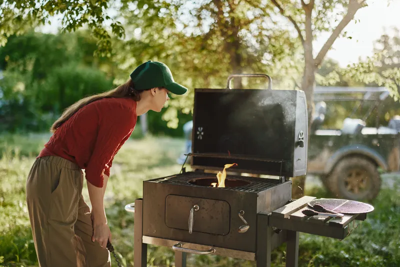 Young woman grilling outdoors in a sunny setting, wearing a red sweater and green cap, enjoying summer BBQ with friends in nature