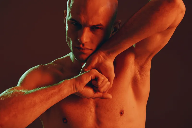 Strong man posing dramatically with an intense expression, showcasing his toned physique and powerful presence. The lighting enhances shadows and highlights.