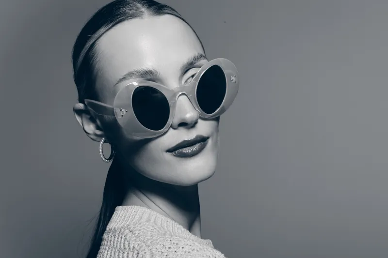 Portrait of a white woman with light skin wearing round sunglasses and hoop earrings in a studio shot.