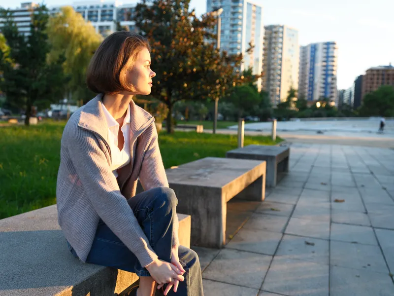 A contemplative woman sits on a concrete bench in an urban park during golden hour. She gazes into the distance, creating a calm, thoughtful, and reflective mood.