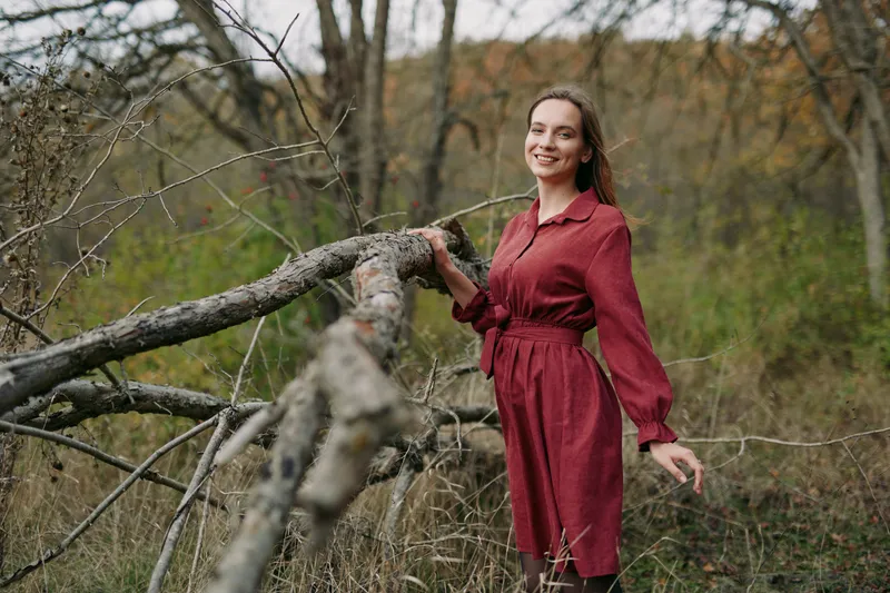 An authentic portrait of a pregnant woman in a red dress beside a weathered log, capturing natural light, relaxed pose, and genuine emotion in a calm autumn landscape.
