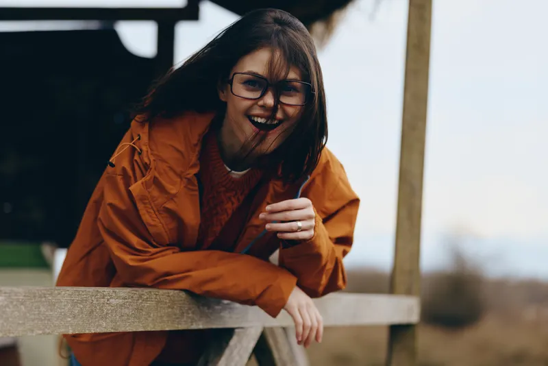 Woman lifestyle outdoors, laughing in orange jacket and glasses, leaning on wooden railing with mug in hand, autumn daylight, casual street moment, warm cheerful mood, gentle breeze