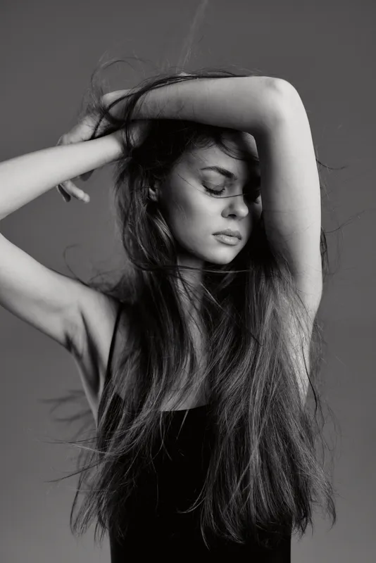 Black and white portrait of a serene woman with long hair raising her arms, expressing calmness and introspection in soft lighting.
