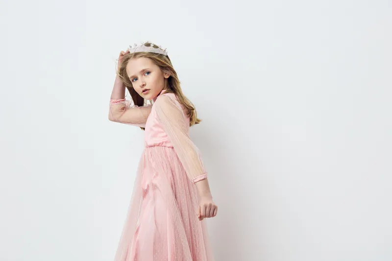 Young girl in a pink dress and crown, showcasing elegance and confidence against a soft white background, perfect for children s fashion and celebrations