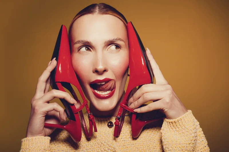 Young woman with red high heels holds shoes near face and looks sideways with playful expression. Studio portrait on yellow background with warm lighting and casual sweater outfit.