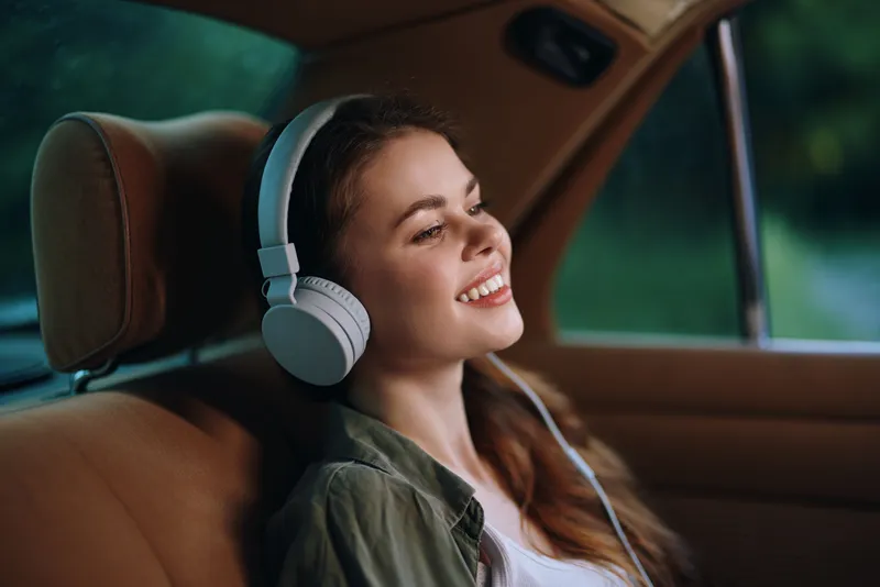 Young woman enjoying music in a car, wearing headphones, smiling with a relaxed expression, summer vibes and a green backdrop, perfect for lifestyle concepts.