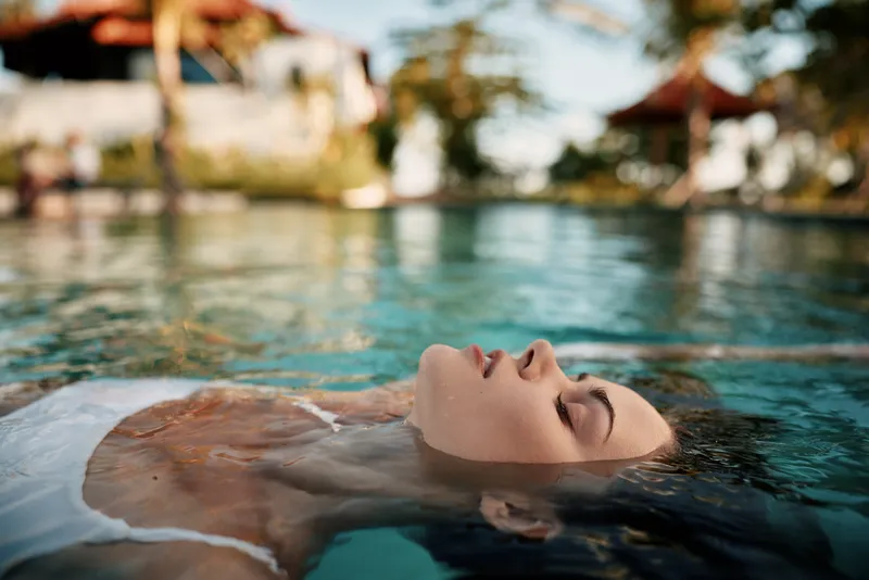 Woman floating in a pool under bright sun, eyes closed, serene expression. Close-up of clear water showing relaxation, wellness, and summer leisure. Gentle ripples and reflections create a tranquil