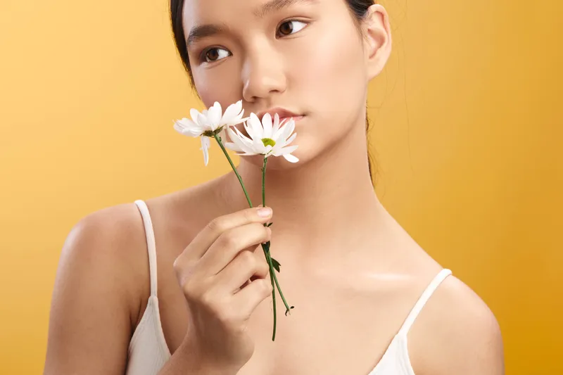 Asian woman gently holds a white flower near her lips, calm studio portrait against a warm yellow backdrop, conveying serenity, fragrance, and feminine beauty in a soft, natural moment.