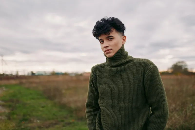 Young man in a thick green turtleneck stands in an open field, captured as a natural, unposed portrait that highlights real moments and a believable, true-to-life presence. (159 characters)