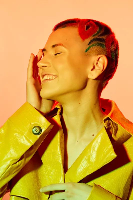 Young woman with creative red patterned buzz cut hairstyle wearing a bright yellow jacket smiling with closed eyes on warm peach background.