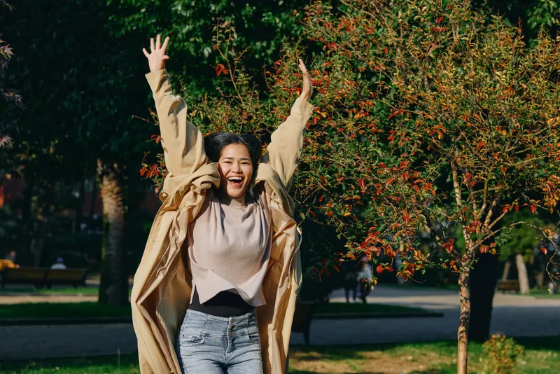 Joyful woman in a beige coat celebrates outdoor in a park, arms raised, celebration of happiness, casual attire, vibrant trees in the background, lively moment with sunlight