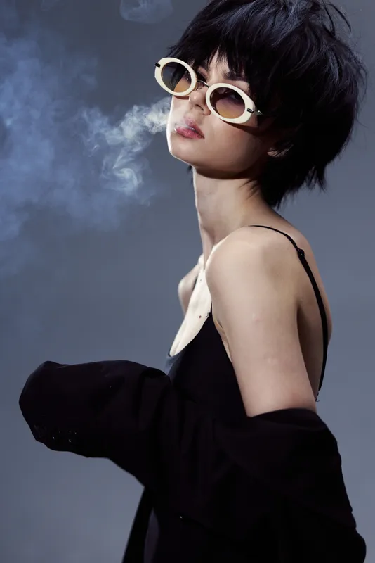 Stylish woman wearing sunglasses and black outfit exhaling smoke with moody lighting and dark background, conveying mystery and confidence.