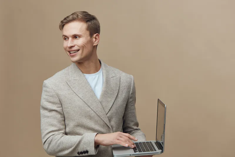 Smiling man in light blazer holding laptop in hand, looking away, wearing casual white t-shirt, standing against plain beige background, studio portrait, people lifestyle concept