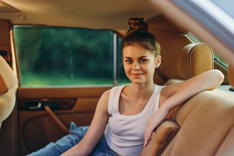 Young woman relaxing in a vintage car interior, wearing a casual white tank top and denim jeans, smiling with a calm expression, showcasing a serene lifestyle.