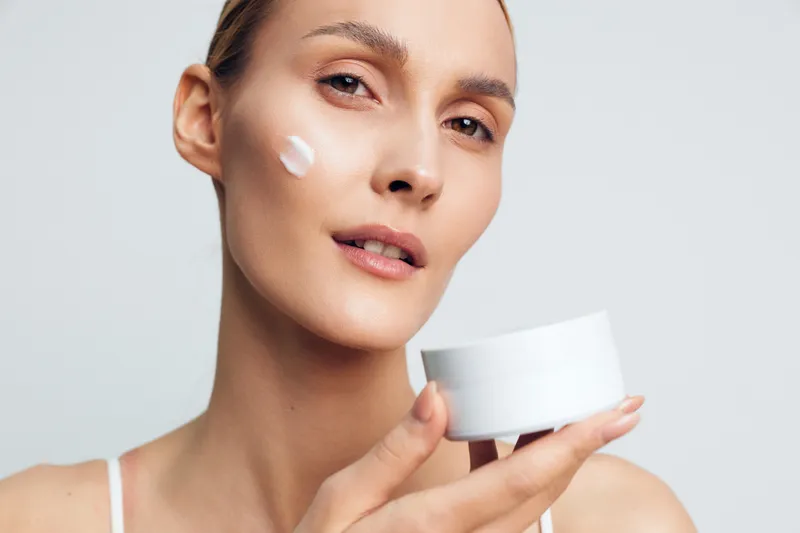 Young woman with clear skin holding white cream jar applying moisturizer on cheek, natural beauty skincare routine, soft lighting, neutral background.