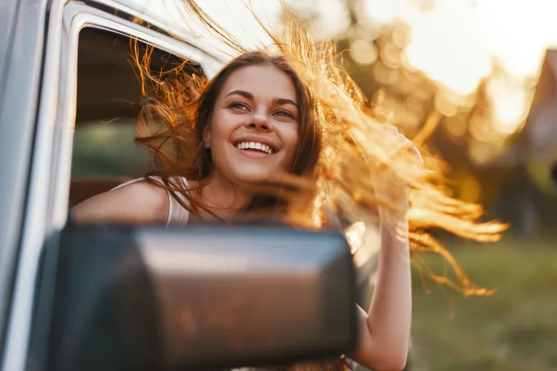 Happy young woman enjoying a road trip with her hair flowing in the breeze, captured during sunset in a natural outdoor setting.