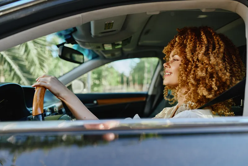 smiling woman driving a car in bright sunlight with natural curly hair and casual beige outfit, enjoying a peaceful moment during daytime.