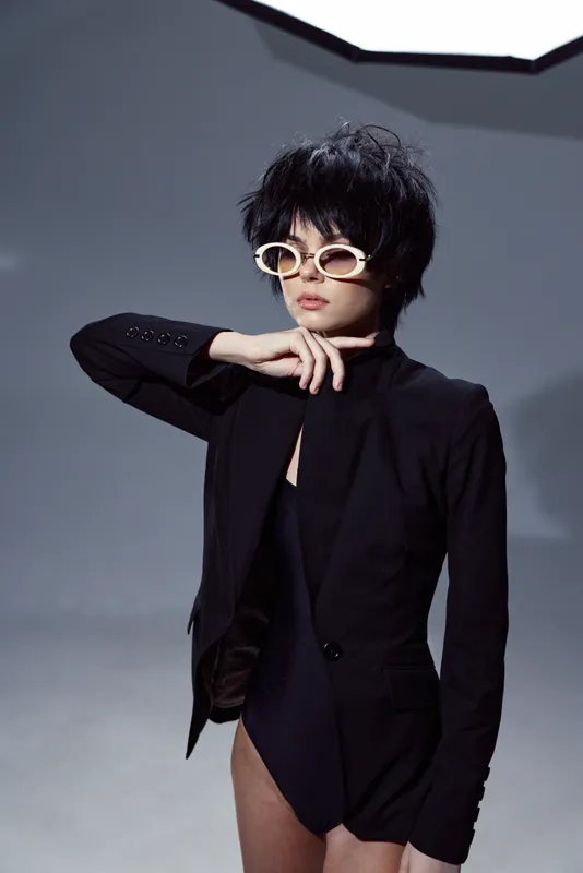 Stylish woman wearing black bodysuit and jacket with trendy sunglasses posing in modern studio lighting.