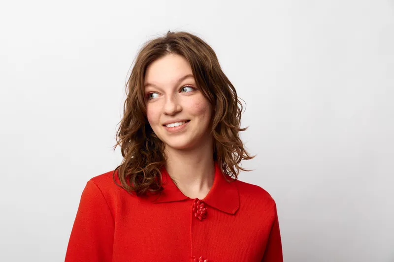 Young woman with curly brown hair wearing red sweater looks sideways with a slight smile. Studio portrait on white background. Concept of casual style and natural beauty.