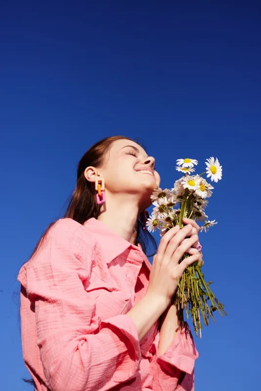 Young woman joyfully holding a bouquet of daisies against a bright blue sky in a pink blouse, symbolizing happiness and summer vibes