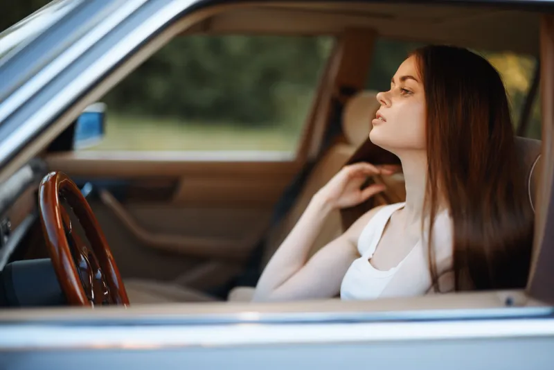 Young woman in a car, lost in thought, with long hair and a serene expression, reflecting on life during a sunny day in nature.