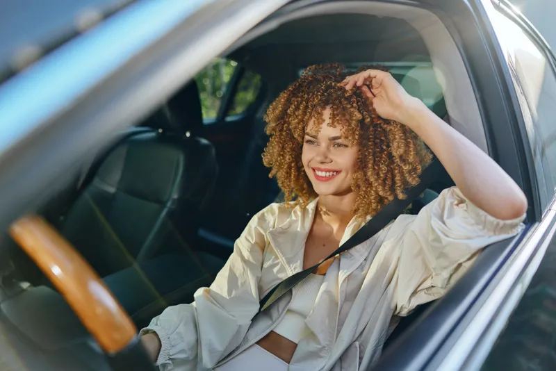 Smiling woman with curly hair wearing casual white clothes, sitting in car with seatbelt fastened, enjoying a sunny day and relaxed mood inside vehicle.